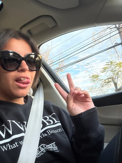 A person, appearing to be female, is depicted in a close-up, chest-up selfie taken from within a vehicle. The subject wears dark, rectangular sunglasses, which obscure the eyes, and a black long-sleeved sweatshirt featuring white text that reads "What w...", "Olivia Benson", and "LAW & ORDER SPECIAL VICTIMS UNIT". A light-colored seatbelt diagonally traverses the subject's chest. The subject's mouth is open with the tongue slightly extended, and their right hand displays a two-fingered "peace" gesture. The background, visible through the vehicle's window, shows a daytime sky with scattered clouds, bare deciduous tree branches, some green foliage, and multiple overhead utility wires. While the environment is consistent with a typical street scene in a city such as Princeton, United States, specific landmarks to definitively identify the precise location within Princeton are not present.