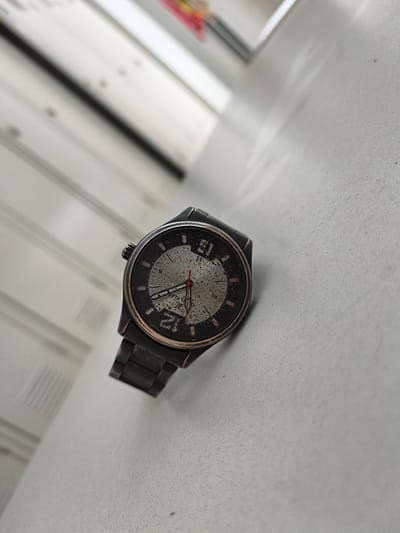 A dark, metal-banded wristwatch is angled on a textured, light gray surface. The watch face is circular with a black bezel and a speckled, grayish-white dial. Red markings indicate the hours, and slender black hands point to approximately 12:40, with a thin red second hand. The watchband is also dark metal. In the background, a blurred, white, slatted surface is visible, with diagonal lines and subtle shadows suggesting blinds or louvers. To the upper right, a small sliver of brightly colored objects, possibly books or toys, is out of focus. The overall scene is a close-up, shallow-depth-of-field shot. The location is Volevčice, Czechia.