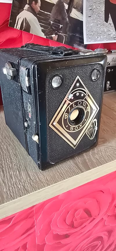 A black Bilora Box camera sits on a wooden surface in the foreground, angled slightly to the right. The camera has a textured body and a metallic diamond-shaped emblem on the front that reads "BILORA BOX" in gold lettering. Two circular viewing lenses are positioned above the emblem. A worn, black strap is attached to the top of the camera. To the left of the camera, there is a silver-colored knob and a latch mechanism. In the background, a photograph is partially visible, showing two individuals. One individual, on the left, is wearing a light-colored jacket over a darker garment and is holding a camera. The second individual, to the right, is mostly obscured. The background is a blurry mix of textures and colors. The scene appears to be indoors, likely on a shelf or table, with a bright pink, rose-patterned fabric in the lower portion of the image. The location is not identifiable from the image itself, and therefore, its presence in Gostagayevskaya, Russia cannot be confirmed.