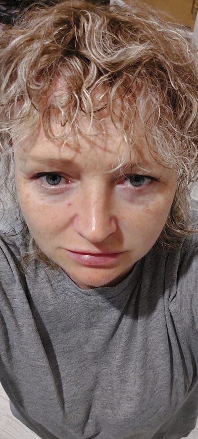The image is a close-up, frontal selfie of a middle-aged white woman with curly, blonde hair. She is wearing a grey t-shirt. Her face is the primary subject, with her blue eyes looking directly at the camera. She has freckles across her nose and cheeks, and there is a small red mark on her right cheekbone. Her lips are slightly parted. The lighting suggests an indoor setting, likely a home environment, and the background is indistinct. There is no discernible location in the city of Зелёный, Russia depicted in this image.