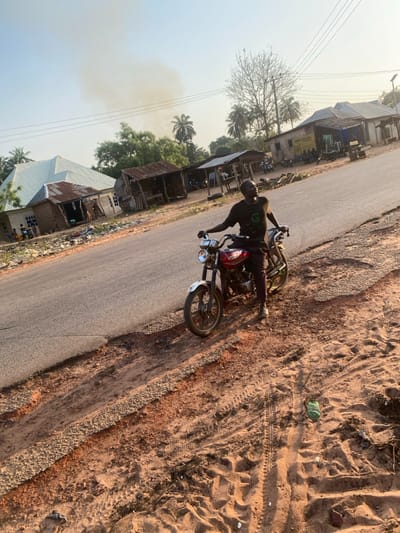 A man wearing a helmet is seated on a motorcycle with a red fuel tank, stopped on a paved road. To the left are buildings with corrugated metal roofs. Beyond the buildings are trees and a hazy sky. The road extends into the distance, while to the right of the motorcycle is a sandy area with tire tracks. A piece of green trash is visible in the sand.