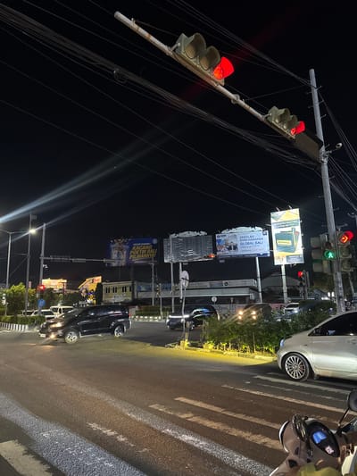 This is a vertical, nighttime, outdoor shot of a busy intersection in Denpasar Barat, Indonesia. The scene is dominated by traffic signals, overhead wires, and illuminated billboards. A white car is parked on the side of the road near a crosswalk, and a white scooter is also visible. The traffic lights are red, indicating that traffic is stopped. The billboards advertise various products and services, including "Kacang Poetri Bali," a brand of peanuts, and "Sekolah Dian Harapan Global Bali," a school opening in July 2027. The overall impression is one of a typical urban night scene in Indonesia, with traffic, advertising, and the general hum of city life. The lighting is artificial, casting long shadows and highlighting the illuminated elements of the scene. The weather appears to be clear, as there are no signs of rain or fog.