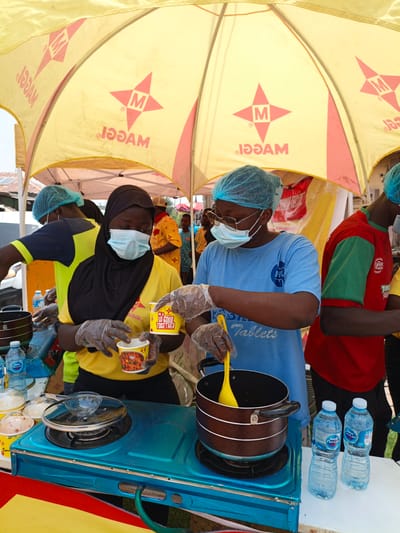 In Tarauni, Nigeria, two young women, wearing protective hairnets and face masks, are preparing food under a yellow Maggi branded umbrella. The woman on the right, dressed in a blue t-shirt, is stirring a pot with a yellow spoon. The woman on the left, wearing a yellow shirt and black hijab, holds two Maggi instant noodle cups, one of which is open, revealing the noodles inside. A blue portable stove sits on a table, with water bottles, an empty pot, and other food preparation items around it. The scene appears to be an outdoor food stall or promotional event.