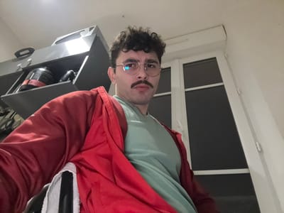 A person with glasses and a mustache looks towards the camera. They wear a red jacket over a green shirt. A window is in the background. A black cabinet with kitchen appliances is also visible. The image is rotated counter-clockwise by 90 degrees.