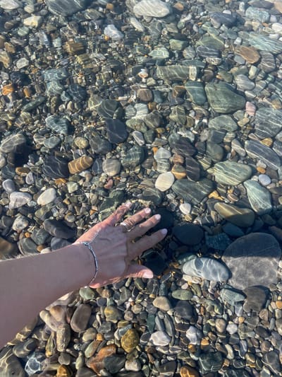 The image captures an overhead view of a person's hand submerged in clear, shallow water over a bed of diverse pebbles and rocks. The hand, wearing a silver chain bracelet and a ring on the fourth finger, is positioned towards the bottom right of the frame, reaching into the water.

The scene showcases a natural setting where the water is exceptionally transparent, allowing for a detailed view of the stones beneath. These pebbles and rocks vary greatly in size, shape, and color, ranging from dark grays, blacks, and greens to lighter browns, whites, and pale blues. Sunlight penetrates the water, creating striking patterns of light (caustics) on the submerged surfaces of the stones, indicating gentle water movement. These patterns appear as shimmering, wavy streaks across the rocks.

The activity depicted is the simple act of a hand interacting with the clear water and its stony bed. The bright illumination suggests it is daytime, likely a sunny day. The clear water and smooth, varied pebbles hint at a clean, natural body of water such as a river, lake, or sea shore, located in Bolshoy Sochi, Russia. There is no visible text in the media file.