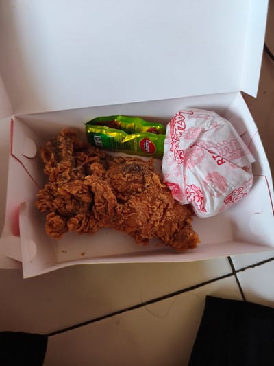 An open white cardboard takeout box containing food items is depicted, resting on a light-colored tiled floor with dark grout lines. Inside the box, a large, golden-brown, breaded piece of fried chicken is positioned on the left. To its right, two small green foil packets, identifiable as "ABC Lazatto" and "HEINZ TOP" chili sauce, are present. Adjacent to the sauce packets, a white paper-wrapped item with red text, likely a burger from "LAZATTO Chicken & Burger," is visible. A dark, possibly black, fabric object is partially seen in the lower right corner of the frame. No individuals are present. The scene is located in the city of Muka, Indonesia.