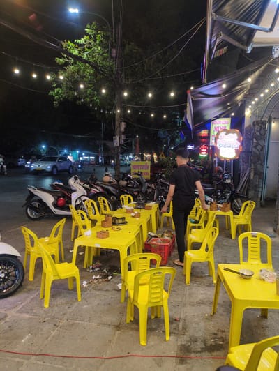 The image shows an outdoor dining area in Tuy Hòa, Vietnam, at night. String lights illuminate the scene. Multiple yellow plastic tables and chairs are arranged on the sidewalk. A person wearing black clothing stands amidst the tables, potentially attending to them. Several parked motorbikes are visible, lining the street adjacent to the dining area. A storefront with illuminated signs in Vietnamese is partially visible in the background. The pavement is concrete.