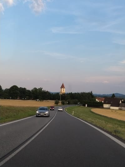 The image captures a view of a rural road in the vicinity of Strakonice, Czechia. The road curves gently to the left, with fields of yellowing crops on either side. In the distance, a prominent church tower with a red roof rises above a line of trees. Several houses with tiled roofs are visible further along the road, suggesting a small settlement. A silver car is the closest vehicle to the camera, followed by a red car and a white car. The sky is a pale blue with wispy clouds. The scene appears to be set in late afternoon or early evening, with soft, diffused light.