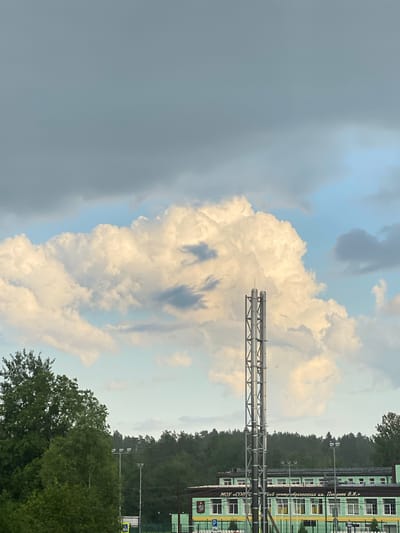 The image depicts a cloudy sky over a forest and a building in Toksovo, Russia. In the foreground, a large, fluffy white cloud dominates the center of the frame, with darker clouds on the left and lighter ones on the right. Jutting out from the right side of the image are two parallel metal pipes, supported by a metal frame. These pipes appear to be part of an industrial or infrastructure setup.
Beyond the pipes, a dense line of trees stretches across the horizon, indicating a forested area. Partially visible behind the trees is a multi-story building with a light green facade and numerous windows, some of which are illuminated. The building's architecture suggests it could be a school, administrative building, or similar public facility. There are signs visible on the building, with some text in Russian. One sign reads "МОУ - СОШ" (which translates to Municipal Public General Education Institution - Secondary School) and another reads "ский центр образования им. П.А. Полякова В.Я." (which translates to educational center named after P.A. Polyakov V.Ya.). There is also a blue sign with a white "P" symbol, indicating parking.
The weather appears to be overcast, with a mix of dark and bright clouds suggesting a dynamic sky, possibly transitioning between rain and clear spells. The presence of lush green trees indicates it is likely summer or late spring. The overall impression is one of an overcast day in a semi-urban or suburban area with natural elements. The scene does not appear to feature any people or specific events.