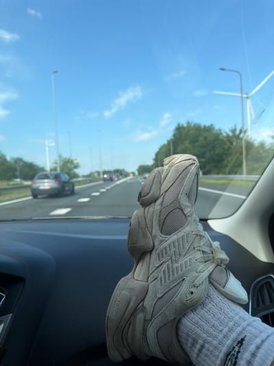 The image shows a close-up view of a person's foot wearing a light-colored sneaker and sock, resting on the dashboard of a car. The car is on a highway, with other vehicles visible in the distance. The sky is blue with some clouds, and trees line the side of the road. Streetlights and a wind turbine are also visible. The scene appears to be captured from inside the car, looking forward. The presence of streetlights and a wind turbine suggests the location might be in the Netherlands, possibly near Breda. The overall impression is that of a journey on a highway under a clear sky.