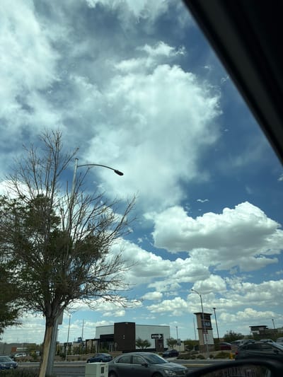 The image, taken from inside a car, shows a view of a street in Las Vegas, United States. The sky is filled with scattered white clouds against a blue backdrop. A leafless tree stands in the foreground, its branches reaching towards the sky. Streetlights are visible alongside the road. In the background, buildings and parked cars suggest a commercial area. The perspective indicates the image was captured while the car was stationary or moving slowly. A black portion of the car's interior frame is visible in the upper left corner. A shopping center sign for ALDI is visible in the distance.