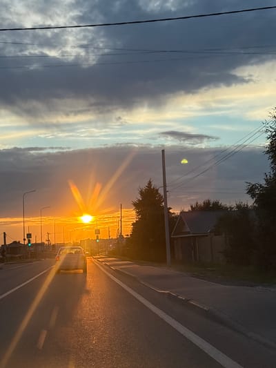 The image depicts a road scene in Kuyuki, Russia, during a sunset. The sun, a brilliant orb with radiating rays, is low on the horizon, casting a warm, golden light across the asphalt road. A white line marks the center of the road, and the surface is slightly reflective from the sunset.

A light-colored car is visible on the right side of the road, heading away from the viewer. Traffic lights, some showing green, are positioned along the roadside, indicating an urban or suburban environment. Streetlights also line the road.

To the left of the road, the sky is a dramatic mix of dark, heavy clouds and lighter, illuminated clouds. The setting sun creates a gradient of colors in the sky, from bright orange and yellow near the horizon to softer blues and grays higher up. In the distance, there are hints of buildings and trees, suggesting a populated area. The overall mood is tranquil, illuminated by the warm glow of the setting sun. The time of day is clearly sunset, and the weather appears clear with some cloud cover.