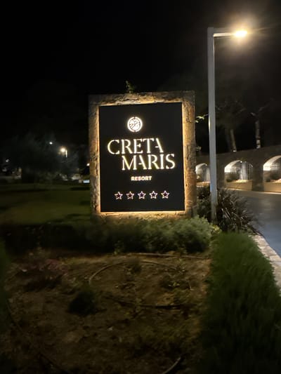 This is a night-time, eye-level, medium shot of a illuminated sign for "CRETA MARIS RESORT" in the city of Chersonisos Municipal Unit, Greece. The sign is framed by a stone border and features the resort's logo at the top, followed by the name of the resort in large, capital letters. Below the name, the word "RESORT" is written in smaller letters, and there are five white stars displayed horizontally. The sign is positioned in front of lush green bushes and a landscaped area, with a paved walkway visible to the right. In the background, there are arched structures with lighting, suggesting the resort's architecture. A street lamp with a bright light is situated to the right of the sign, casting a glow on the scene. The overall environment appears to be well-maintained and inviting, typical of a resort setting.