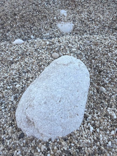 The image shows a beach scene with a large, white stone resting on a bed of small, smooth pebbles. The pebbles are primarily light gray and beige in color, varying in tone and size. The large stone is irregularly shaped and textured. The location is likely the Municipal Unit of Lefkada, Greece, as the geology and type of beach stones is typical of the region. There are no people visible.