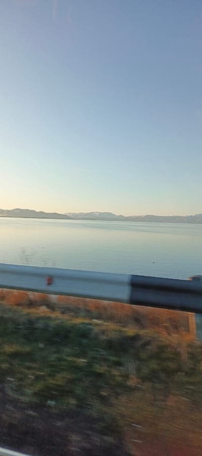 The image captures a serene landscape from a moving vehicle, featuring a vast body of water framed by distant mountains under a clear sky.

In the foreground, a metallic guardrail runs horizontally across the bottom half of the frame, indicating the proximity of a road or highway to the water's edge. Below the guardrail, blurred green and brown foliage suggests motion, as if the photograph was taken from a car or bus in transit.

The middle ground is dominated by calm, reflective water, appearing light blue-green under the soft light. The water surface shows subtle ripples and reflections, with a small, dark, indistinct object barely visible in the distance on the water.

In the background, a range of mountains stretches across the horizon, some peaks distinctly capped with snow, adding to the tranquil and somewhat cool atmosphere. The mountains recede into a gentle haze, enhancing the sense of depth.

The sky above is clear and light blue, transitioning to a paler, almost white, hue near the horizon, indicative of either early morning or late afternoon light. There are no visible clouds. The overall impression is one of peace and expansive natural beauty. No people or discernible text are visible in the image. Given the location context of Berdkunk, Armenia, the body of water is likely Lake Sevan.