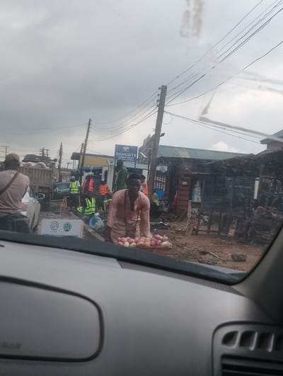 The image captures a street scene in Iju, Nigeria, under an overcast sky, suggesting a cloudy or potentially rainy day. The foreground is dominated by the interior of a vehicle, with the dashboard and windshield visible, indicating the photo was taken from a car.
In the midground, a man in a light brown, traditional garment stands in front of a cart overflowing with onions. He appears to be a vendor, showcasing his produce. Behind him, a bustling street scene unfolds. Several individuals wearing high-visibility vests are scattered around, possibly involved in construction or road work, given the presence of a truck loaded with cement bags. A sign for "DANGOTE CEMENT DEPOT" is clearly visible, confirming the location's industrial or commercial nature.
The environment is unpaved and appears somewhat disorganized, with loose dirt and debris on the ground, characteristic of many developing areas. A few small shops and stalls line the street, suggesting a market or commercial zone. The overall atmosphere is one of everyday activity, with people engaged in their work and commerce amidst the daily hustle and bustle. The lighting is muted due to the cloudy weather, casting a soft, diffused light over the scene.