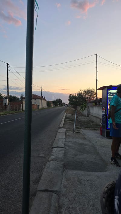 The image depicts a street scene in Mirovo, Bulgaria, at dusk. A dark green pole dominates the foreground, obscuring a portion of the view. The street extends into the distance, lined with houses and trees on either side. Power lines crisscross the sky, supported by utility poles. On the right side of the street, a Pepsi vending machine stands next to a concrete wall. A person wearing a teal shirt and blue shorts is standing near the vending machine. The sky is filled with soft pink and pale blue clouds, indicating the setting sun. The ground in the foreground is a mixture of asphalt and concrete, with a concrete curb running along the side of the road.