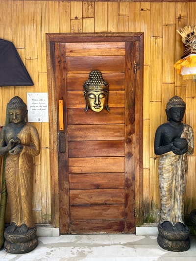 The image captures a scene at a location in Singakerta, Indonesia, likely a bathroom or spa area, as indicated by a sign requesting patrons to shower before using the bath tubs and pool. The scene is framed by bamboo walls, with a dark wooden door in the center. A brass Buddha head is mounted on the door. Two reclining Buddha statues, painted black and gold, are positioned above and below the door, adding to the spiritual ambiance. The floor is tiled in white and the overall atmosphere is that of a tranquil and contemplative space.