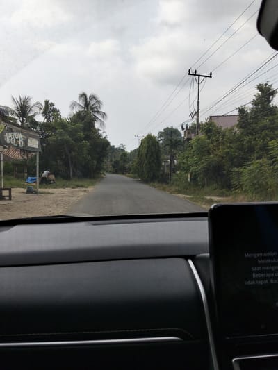 The image captures a view from inside a vehicle, looking out onto a two-lane paved road in Blang Mee, Indonesia. The sky is overcast, with visible clouds. On the left side of the road, there's a roadside stall or building with a sign that is partially visible and reads "NATION BLD." Adjacent to this structure, a person is observed standing next to a large basket, seemingly engaged in some activity. Several blue tarps are laid out on the ground. Various trees, including palm trees, line the road. Further down the road, a utility pole with wires and a transformer is visible. On the right side of the road, houses and more trees can be seen. The interior of the vehicle shows a dashboard with a digital display screen, which has text in what appears to be Indonesian.