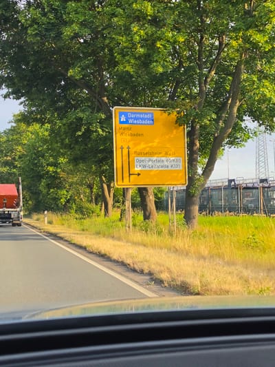 The image shows a German road sign in Rüsselsheim am Main. The sign is yellow and rectangular with blue text and symbols. It indicates directions to Darmstadt and Wiesbaden, with the Autobahn symbol visible. Below this, it directs towards Mainz and Wiesbaden, with the number 43 in a red square. Further down, it indicates a route to Rüsselsheim-West, with specific destinations of "Opel-Portale 60/K80" and "LKW-Leitstelle K131" and a left-pointing arrow. The sign is positioned next to a tree-lined road with dry grass on the side. In the background, there are industrial structures and a transmission tower. A red truck is visible on the left side of the road. The perspective is from inside a vehicle, looking out.