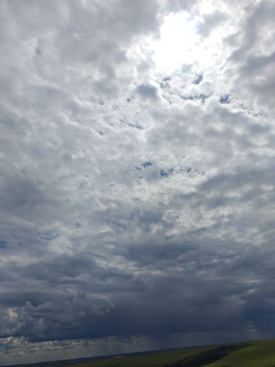This is an overcast sky over a rural landscape, likely in or near Colonița, Moldova. The sky is filled with a variety of clouds, ranging from thin, wispy formations to darker, more ominous storm clouds in the lower portion of the image. Patches of blue sky are visible through the clouds in the upper half of the frame, and the sun is partially obscured, creating a bright glow. The landscape below consists of rolling green hills, with some darker patches that could be forests or fields. A hint of distant buildings or structures can be seen along the horizon line. There are no people or animals visible in the image. The overall impression is one of a dynamic and changeable weather system.