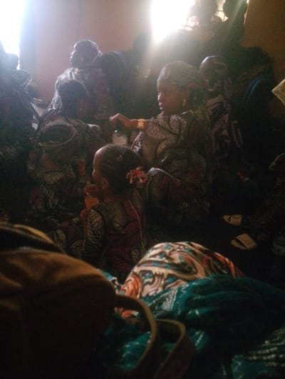 A group of people, likely women and children, are gathered indoors. They are dressed in patterned fabrics, some wearing head coverings. A girl in the foreground has flowers in her hair. The scene is brightly lit with a light source behind them. The lighting is strong and creating shadows. The image's foreground is blurred.