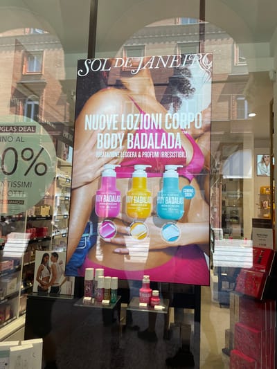 The image shows a storefront window of a cosmetics and perfume store, likely in Bologna, Italy, based on architectural reflections. The store displays advertisements for brands like Sol de Janeiro, Armani, Dior, and Moschino, as well as numerous perfume and cosmetic products on shelves visible through the glass. There's also an advertisement indicating a "Douglas Deal" of 0% off "Santissimi Prodotti" or "Holy Products" of "Diori Marchi" brands.
The main focus is a poster featuring Sol de Janeiro's "Body Badalada" lotions, advertising them as lightweight and irresistible. The products are showcased with imagery of a woman, skin, and clothing. Reflections on the glass show buildings across the street. There is a Moschino advertisement with a price of €55,99. The scene appears to be during daylight, given the brightness.