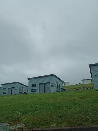 The image shows a cluster of modern, light blue cabins situated on a grassy hillside in Doirí Beaga, Ireland. The cabins have dark gray roofs and dark-framed windows, some of which are large glass doors. One cabin on the left has text on its side that appears to read "Inis Meain". A picnic table is visible in front of this cabin. Further up the hill, more cabins or buildings can be seen, one with white walls and a dark roof. The sky is overcast and gray, suggesting cloudy weather. The foreground consists of a vibrant green lawn dotted with small white flowers. Rocks and a dark border are visible at the very bottom of the image, possibly indicating a pathway or edge of a property. There are no people visible in the image, and the cabins appear unoccupied. The scene is static and depicts a residential or holiday accommodation setting.