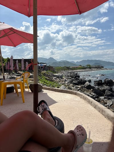 The image captures a sunny day at a coastal location in Vĩnh Lương, Vietnam. The foreground shows a person's legs resting, one with a black and white sandal and the other with a pink sandal. Next to the pink sandal is a clear plastic cup with a straw containing a yellow beverage. In the midground, there is a yellow plastic table and chair set, with several folded pink umbrellas in the background. Beyond the seating area, a rocky coastline meets the ocean, with gentle waves and distant mountains visible under a partly cloudy sky. A few buildings and vegetation are scattered along the coast. The scene appears to be an outdoor seating area at a beachside establishment.