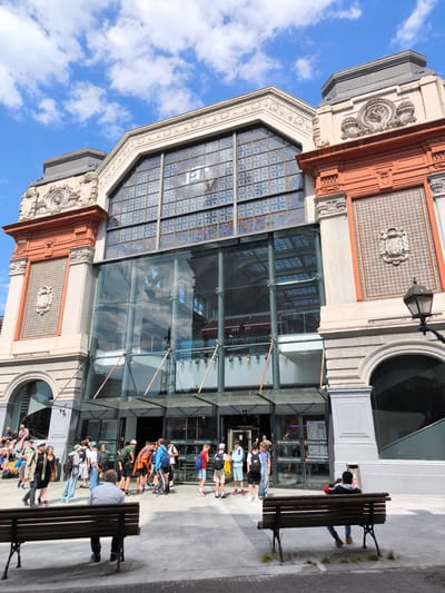 The image shows the exterior of the Mercado de la Ribera in Bilbao, Spain. The market is a large Art Deco building with a prominent stained-glass window above a modern glass entrance. A group of people, many of whom appear to be tourists with backpacks, are gathered near the entrance. Some are sitting on benches in the foreground. The sky is blue with white clouds.