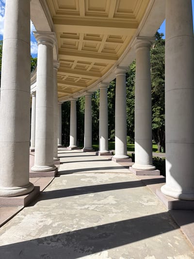 The image shows a colonnade with a curved roof supported by numerous large, light-colored columns. The columns are cylindrical and have distinct segmented bases. The roof has a coffered ceiling painted in a pale yellow. Sunlight casts strong shadows across the paved walkway in front of the colonnade, creating a pattern of light and dark. The background consists of lush green trees and a clear blue sky with scattered white clouds. The scene appears to be outdoors in a park-like setting, possibly a historic site or a public garden. The location is identified as the посёлок дачного хозяйства «Архангельское», Russia, suggesting it is part of the Arkhangelskoye estate. There are no people visible in the image. The architectural style suggests a classical influence, and the overall impression is one of grandeur and serenity.
