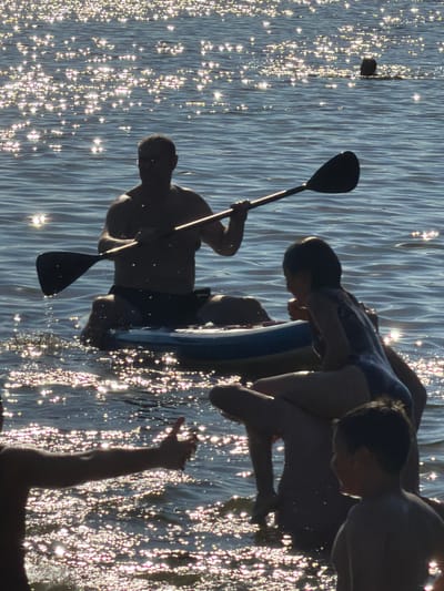 In Malakhovka, Russia, on a sun-drenched day, a man is paddleboarding in the shimmering water, holding a paddle. He is accompanied by a young girl, who is also on the paddleboard with him, and a younger boy who is standing in the water nearby. The water is clear and blue, with sunlight reflecting off its surface, creating a dazzling display of sparkles. The scene is one of leisure and outdoor recreation, with the individuals enjoying the warm weather and the refreshing water. The overall impression is one of a peaceful and happy day spent at the lake.