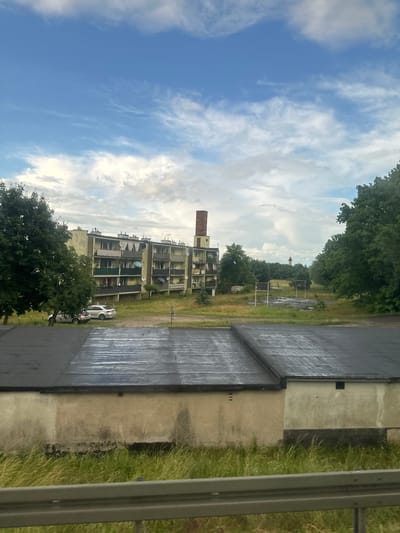 The image captures a view of a residential building and its surroundings in Połupin, Poland. In the foreground, two flat-roofed structures with dark, weathered roofs are visible. The walls of these structures are a pale, off-white color, appearing somewhat aged with visible stains and textures. Tall, green grass grows along the base of these buildings, and a gray metal guardrail runs across the bottom of the frame, suggesting the viewpoint is from a road or elevated platform.
Behind these foreground structures, a multi-story residential building dominates the mid-ground. It is painted in light yellow and green hues, featuring numerous balconies with railings. A tall brick chimney stands adjacent to this building. The building appears to be in a state of disrepair or neglect, with some balconies exhibiting clutter. Several cars are parked in front of the building, on a grassy area.
Further in the background, a field with overgrown grass and what appears to be a derelict basketball court with rusted hoops is present. The landscape is dotted with mature trees, forming a dense treeline. In the far distance, a church tower with a steeple is faintly visible through the trees, under a sky filled with scattered white and grey clouds against a blue backdrop. The overall impression is that of a somewhat run-down urban or semi-urban environment.