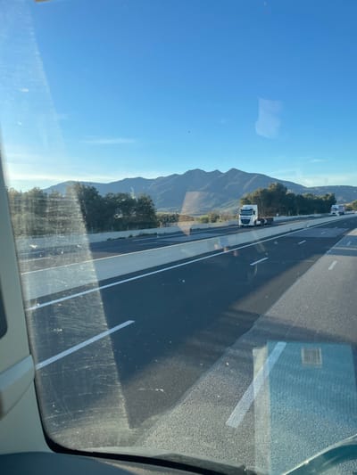 The image captures a daytime highway scene, viewed from inside a vehicle through its windshield, situated near Tresserre, France. The upper portion of the frame is dominated by a clear, bright blue sky with minimal cloud cover. A range of dark green, rugged mountains stretches across the horizon, with sporadic trees visible in the mid-ground between the mountains and the roadway. The foreground features a multi-lane highway constructed of dark asphalt, delineated by white dashed and solid lane markings. A light grey concrete median barrier separates the directions of traffic, casting distinct shadows onto the road surface, indicative of strong, direct sunlight. In the lanes of opposing traffic, a white DAF semi-truck tractor, without a trailer, is visible in the leftmost lane, traveling away from the perspective of the camera. Further down the opposing road, another white semi-truck is partially discernible. The vehicle's windshield, framing the lower-left portion of the image, exhibits reflections and minor surface imperfections.