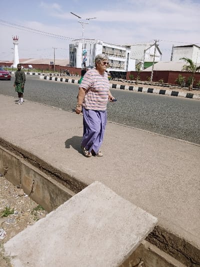 The image depicts an outdoor urban environment in Communauté rurale de Payar, Senegal, under clear, bright daylight. In the mid-right, an elderly Caucasian female, with short gray hair, sunglasses, a horizontally striped t-shirt (white, red, light blue), purple loose-fitting trousers, and light-colored closed-toe shoes, walks on a concrete sidewalk, holding a dark object in her right hand. Her left hand is at her side, wearing a bracelet. In the mid-left, a younger dark-skinned male, wearing a dark long-sleeved shirt, green patterned knee-length shorts, and white sneakers, stands on the concrete sidewalk adjacent to an asphalt road. A red sedan is partially visible on the road behind him. The road is bordered by black and white striped curbs. In the background, a multi-story white building with balconies is prominent, alongside other structures with reddish-brown walls and a distinct tall white cylindrical tower topped with a red crown-like structure. A solar-powered streetlight is visible above the background buildings. A concrete-lined drainage channel runs along the sidewalk edge in the foreground, partially covered by a concrete slab, with visible debris within the channel.