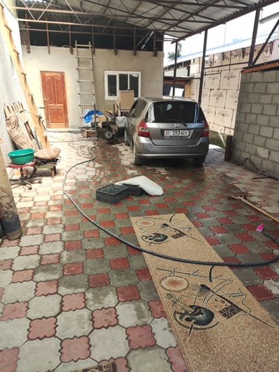 The image captures a partially covered outdoor courtyard or workspace in Chong-Aryk, Kyrgyzstan. A grey Honda Fit hatchback is parked prominently, facing away from the viewer, its license plate reading "01 193 ACV". Adjacent to the car is a brick wall and construction materials, including what appears to be insulation panels with Cyrillic lettering. The ground is paved with interlocking concrete tiles in a red and grey pattern, and there are puddles of water, suggesting recent rain or cleaning.
To the left, a building with a wooden door and a window is visible, with a tall wooden ladder leaning against its wall. Various items are scattered around, including a green plastic basin on a red wheeled stool, a coiled hose, two black plastic crates, and a long, patterned rug laid out on the ground. The scene is under a corrugated metal roof supported by metal beams, indicating it might be a carport or an open-sided structure. The overall impression is one of a functional, somewhat cluttered outdoor area, possibly undergoing renovation or in use for storage and work. There are no people visible in the image.