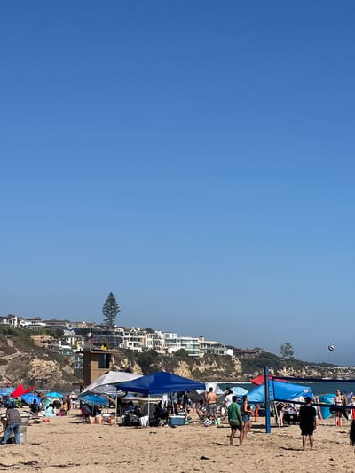 The image depicts a crowded beach scene on a sunny day. A volleyball net is set up in the center of the frame, with several people engaged in a game. Beach umbrellas and tents are scattered across the sand, providing shade for beachgoers. In the background, a lifeguard tower stands on a rocky cliff overlooking the ocean, with houses perched on the hillside above. The city of Newport Beach, California, is known for its beautiful coastline and recreational activities, making it a popular destination for both locals and tourists. The clear blue sky and calm ocean waves suggest ideal beach weather.