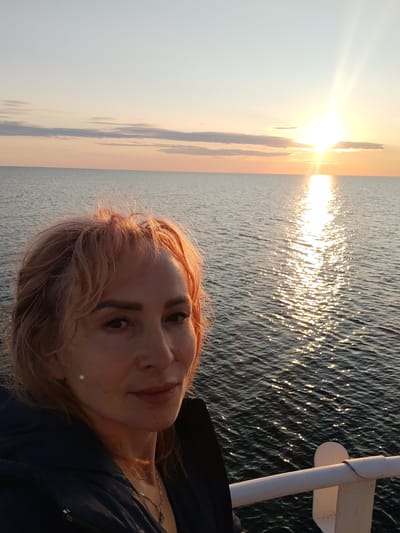 A woman with auburn hair is positioned in the foreground, facing slightly to her left, with the ocean and a sunset in the background. She is wearing a dark jacket and a necklace. The sun is setting over the water, casting a bright reflection. The image appears to be taken from a boat or pier, indicated by a white railing in the lower right corner. The location is described as Kakkarovo, Russia.