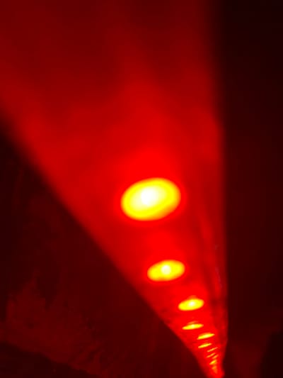 A low-angle, close-up image features a string of red lights with a yellow center. The lights recede into the distance, appearing smaller and closer together. The surface they are on appears dark. The color palette is primarily red and yellow, with the red hue dominating the overall scene. The lighting creates a dramatic effect.