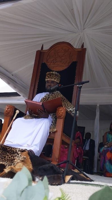 A man wearing a leopard skin headdress and a white robe sits on a throne with a carved wooden back and arms, holding a red book. The throne is under a white fabric canopy. A microphone is mounted on a black stand in front of him. There is a large leopard skin lying on the floor in front of the throne, and a black dog is partially visible on the skin. The man is speaking into the microphone. In the background, there are other people sitting in chairs, one of whom is a man in a suit and tie. The image is likely taken in a large hall or a room with a high ceiling. The location, based on the coordinates (-1.624, 29.258), is Rukoko, Democratic Republic of the Congo.