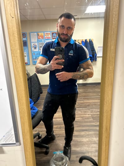 A male individual is depicted taking a mirror selfie within an ALDI staff room, identified as being in Lancaster, United Kingdom. The man has short dark hair, a beard, and visible tattoos on both forearms. He is wearing a dark blue ALDI polo shirt with white and light blue trim, featuring the ALDI logo and text on the chest, along with a nametag on his left side. He also wears black cargo pants and black athletic shoes. His right hand holds a smartphone with a circular grip accessory, pointed towards the mirror. The reflected background reveals a functional staff area with light-colored walls and wood-patterned laminate flooring. On the left wall, several ALDI-branded informational posters are displayed. To the right, a rack holds multiple blue uniform jackets. A portion of a black office chair and a clear plastic jug are visible on the floor in the lower part of the frame.
