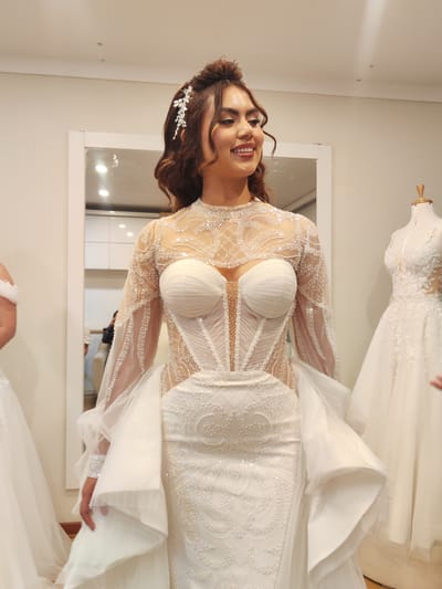 A woman stands in what appears to be a bridal shop in Tacna, Peru, wearing a long-sleeved, white, beaded wedding gown with a corset-style bodice. She is smiling and looking slightly to her right. She wears a silver hair ornament on the left side of her head. The room contains at least two other wedding dresses on mannequins, and there is a mirror behind the woman. The lighting is bright, suggesting an indoor environment.