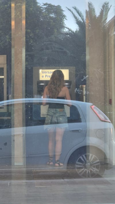 The image, taken in Taranto, Italy, captures a reflection on a glass surface, creating a layered and somewhat distorted view of the surroundings. The primary subject is a young woman seen from the back, leaning into the open driver's side window of a car. She has long, wavy brown hair and is wearing denim shorts and a dark-colored top. Her legs are visible, and she is wearing dark sandals.
Through the car window, a yellow sign with text is visible, but it's partially obscured and the writing is difficult to decipher clearly. However, the word "Versan" can be read on the sign, possibly part of a business name or service. The car itself appears to be a light-colored hatchback, with its rear tire visible through the reflection.
Outside the car, the reflection also shows elements of the environment. A large palm tree frond is prominent on the left side of the image, suggesting a warm climate and possibly a park or landscaped area. Green foliage from trees is also visible, interspersed with the urban reflection. The lighting suggests it is daytime, with the sun casting reflections on the glass. The overall scene is one of a casual, everyday moment, with the woman seemingly interacting with the car or something within it. The reflections create a sense of depth and complexity, blending the interior of the car with the exterior scenery and the viewer's perspective.