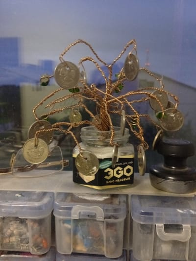 The image shows a decorative tree made of copper wire and coins, placed in a jar. The jar appears to contain some sort of paste or cream with text on it that reads "ЭГО" and "БЛИС МЕДОВЫЙ". To the left of the jar are a pair of glasses, and to the right is a black ink pad. Behind these objects, out of focus, is a window showing a blurry cityscape. Below the shelf holding these items are several clear plastic storage containers filled with various small objects. The scene appears to be indoors, possibly in a workshop or hobbyist's area. The style of the coins and the text on the jar suggest a Russian origin. The overall aesthetic is one of handmade craft and practical organization. The specific location in Nyurba, Russia, cannot be determined from the visual information provided in the image.