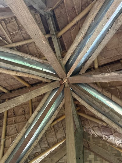 The image presents an upward interior view of a rustic roof structure located in Ban Paem Bok, Thailand. The central element features multiple weathered wooden beams, roughly squared, converging radially towards a central apex secured by visible wooden cross-members. These primary beams extend outwards, supporting an overhead roof composed of layered and woven dried organic material, consistent with thatch or palm fronds, which appears aged and shows some irregularities. Numerous cylindrical bamboo poles are visible, providing additional structural support beneath the thatched material. Integrated within the roof framework, specifically between some of the wooden beams, are sections of reflective, translucent corrugated sheeting, likely clear plastic or thin metal, which reflect ambient light. No individuals, actions, or specific interactions are depicted. The overall construction suggests traditional or vernacular architecture.