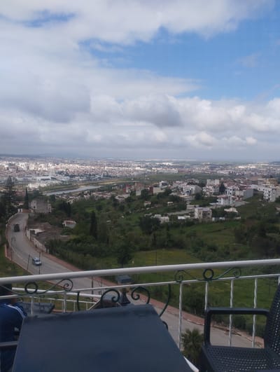 A high-angle daytime view shows part of Tétouan, Morocco. The foreground includes a portion of a balcony with a white railing, a dark-colored table, and a black chair. A person wearing a blue jacket is partially visible. Below the balcony, a winding road is visible, with a light-colored car on it. Beyond the road, green vegetation covers the terrain. A mix of residential buildings is scattered among the greenery. In the distance, the city of Tétouan extends toward the horizon, obscured by clouds. The sky is cloudy with patches of blue.