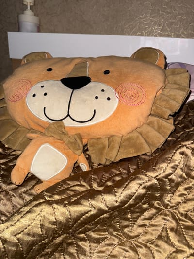 A stuffed toy lion is lying on a quilted, shiny brown blanket. The lion is orange with a brown mane and ears, and has a white face with black dots for eyes and nose, and a stitched smile. It is wearing a small brown bow tie. In the background, a white shelf holds a white pump dispenser. The wall behind the shelf has a textured, beige wallpaper. The image is taken from a slightly elevated angle, looking down at the toy. The scene appears to be indoors, likely in a bedroom. There is no indication of the location being in Novoshakhtinsk, Russia.