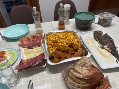 A table laden with food in Γερακινή, Greece, displays various dishes and items. A light blue bowl sits on the left, next to a plate with feta and olive oil. Another plate holds prosciutto, cheese, and pastrami. A central aluminum tray contains french fries and fried chicken pieces. A white plate to the right has a grilled fish. Other items include a container of olives, a jar of pickles, bottles of oil and vinegar, a salt shaker, glasses, forks, and a bag of bread. Two brown chairs are visible in the background. The tablecloth is white with a checkered pattern.