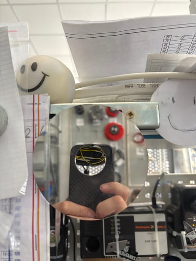 The image depicts a close-up view of a cluttered workspace with various electronic components, wires, and papers. In the foreground, a round, reflective object, likely a mirror or a polished surface, shows a distorted reflection of a hand holding a black object with yellow wires. Behind this, a white object with a drawn smiley face is visible, along with more papers and cables. The background is filled with shelves, electronic equipment, and some labels. The setting appears to be an indoor, possibly a workshop or laboratory, environment. There is no information in the image to indicate the location in Novocherkassk, Russia.