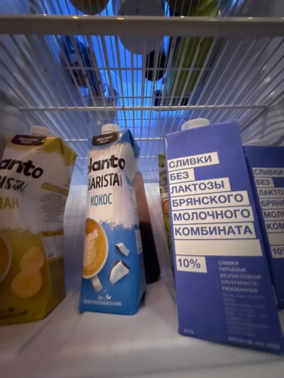 Inside a refrigerator, two cartons of milk-like beverages and another yellow carton are visible on a shelf. The carton on the left is yellow and appears to have text in a dark color, with an image of banana slices. The carton in the middle is white and blue, with "Planto BARISTA KOKOS" written on it. It also features an image of coffee with latte art and coconut pieces. To the right of this, a blue carton with white text reads "СЛИВКИ БЕЗ ЛАКТОЗЫ БРЯНСКОГО МОЛОЧНОГО КОМБИНАТА" (Cream without lactose from Bryansk Dairy Plant) and "10%". Another similar blue carton is partially visible to the far right. The shelves are made of white wire. The overall environment suggests a domestic refrigerator. The location is identified as Veshki, Russia.